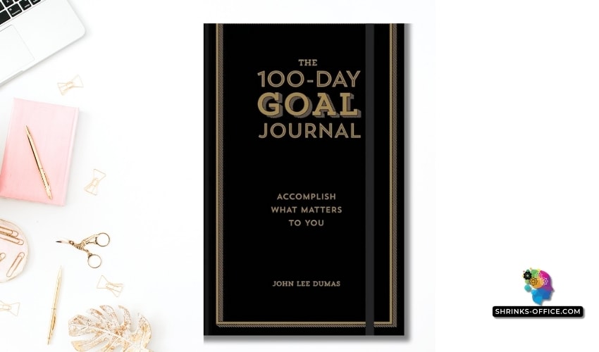 A stylish and minimalist workspace featuring 'The 100-Day Goal Journal' by John Lee Dumas, alongside a laptop and elegant gold stationery.